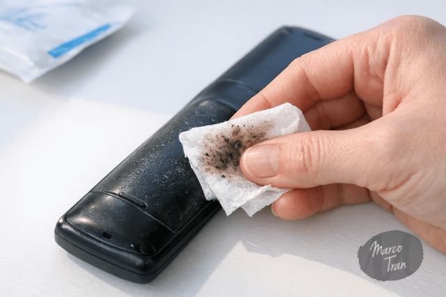 A photorealistic image of a hand using an alcohol wipe to clean the sticky rubber surface of a black remote control on a minimalist white table, with visible residue on the wipe, crisp natural light, realistic detail, not AI generated